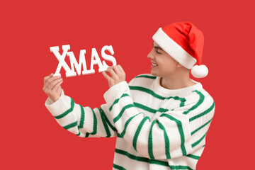 Young man in Christmas hat holding word XMAS on red background