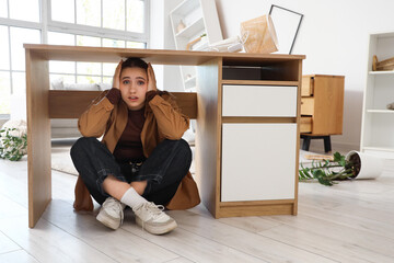 Scared young woman hiding under table at home during earthquake