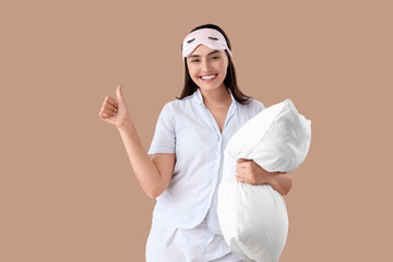 Young woman in striped pajamas with pillow showing thumb-up on beige background