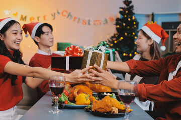 Group of young Asian man and women as friends having fun at a New Year's celebration, holding gift boxes standing by Christmas tree decoration, midnight countdown Party at home with holiday season.