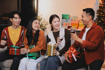 Group of young Asian man and women as friends having fun at a New Year's celebration, holding gift boxes standing by Christmas tree decoration, midnight countdown Party at home with holiday season.