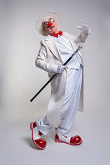 An elderly clown in a white coat, hat and large red patent leather clown boots. Walking stick in hand.