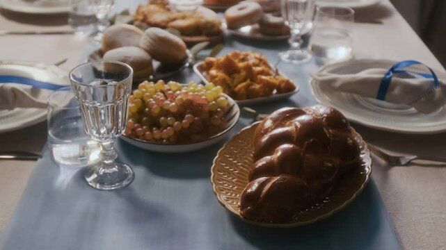 Close up view of festive dining table set with traditional foods like challah, sufganiyah and grapes, elegant glassware, plates and blue-accented napkins on Hanukkah holiday