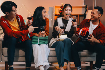 Group of young Asian man and women as friends having fun at a New Year's celebration, holding gift boxes standing by Christmas tree decoration, midnight countdown Party at home with holiday season.