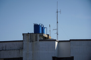 A blue water tank is seen on the rooftop of a building with a television antenna. A rooftop view with water tanks and an antenna bathed in the warm glow of a sunny day.