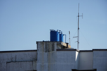 Three blue water tanks on a rooftop, with a tall antenna. The building is made of concrete and has a white paint job. Blue Water Tanks on a Rooftop Background.