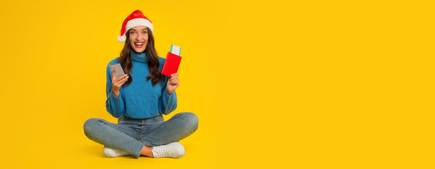 Happy woman in a festive Santa hat sits cross-legged on a vibrant yellow background, holding a phone in one hand and colorful gifts in the other, radiating excitement and holiday spirit, copy space