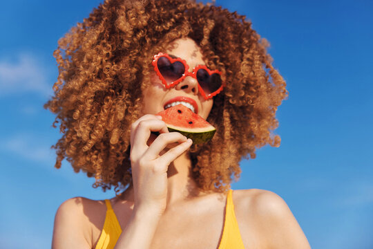 Young woman with curly hair enjoying watermelon on a sunny beach with a clear blue sky Her playful heart shaped sunglasses add a fun touch