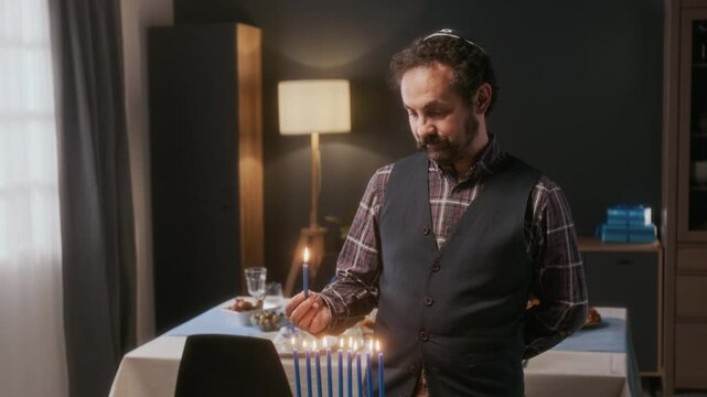 Jewish man wearing kippah lighting menorah candles for Hanukkah celebration in cozy living room, with festive dinner table in background