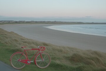 Fototapeta premium A red bicycle sits on a sandy beach with a picturesque view of the ocean and a coastline in the background.