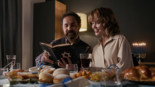 Smiling Jewish couple reading sacred text in prayer book with Star of David at festive table with traditional food and Kiddush wine during Hanukkah celebration at home