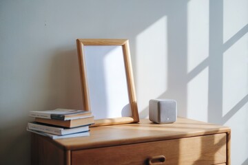 Wooden frame with blank white paper on dresser, paired with books and cube speaker, softly lit by natural light against light wall, creating a peaceful setting