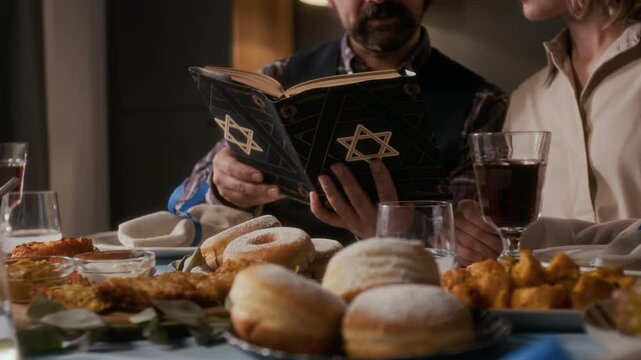 Close up view of Jewish couple sitting at festive table, reading prayers from Siddur book during Hanukkah celebration at home