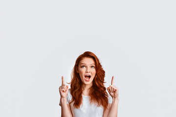 Look Up. Red-Haired Teen Girl Pointing Fingers Upward At Free Space Posing Over White Background. Studio Shot