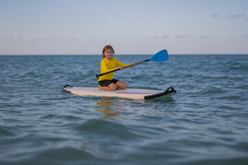 Boy swimming on stand up paddle board. Water sports, active lifestyle. Kid paddling on a paddleboard in the ocean. Child Paddle boarder. Summer Water sport, SUP surfing. Summer beach vacation.