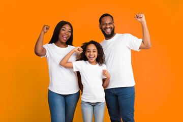 Fototapeta premium Yes. Portrait of excited young black family of three people shaking clenched fists, making winner gesture. Happy African American man, woman and girl celebrating victory together, yellow orange studio