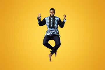 Emotional black guy showing happiness over pink studio background. Smiling african american man in colorful traditional clothes jumping up and posing, raising fists up, expressing emotions