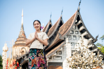 A beautiful Asian woman stands outdoors in a praying posture, with a temple in the background.