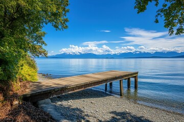 Wooden dock extending into a calm, blue lake with a mountain range in the distance on a sunny day.