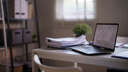 An organized office desk with a laptop, documents, and a small potted plant, set against a background of shelves and window blinds