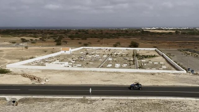 Elevated perspective of Rabil Cemetery on Boa Vista, Cape Verde, set amidst a barren desert with distant ocean views, capturing the stark contrast of the white walls against the rugged surroundings