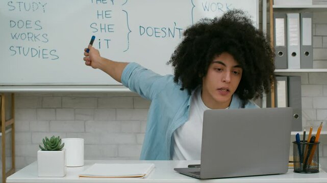A young student with curly hair sits at a desk, focused on a laptop screen while studying English grammar concepts. A whiteboard in the background displays English pronouns and verbs.