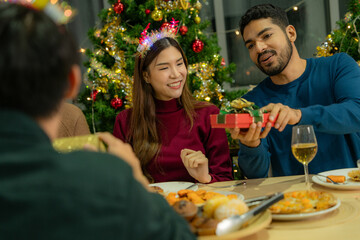 Group of Asian friends enjoying Christmas party for new year celebrate. Friends having fun together at home. men and women opening box gift and present together at home with Christmas tree.