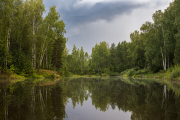 Summer landscape with a forest lake. Evening view of a small lake in the forest. Birches grow along the banks. Trees are reflected in the water. Cloudy weather. Clouds in the sky. Outdoor recreation.