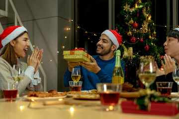 Group of Asian friends enjoying Christmas party for new year celebrate. Friends having fun together at home. men and women opening box gift and present together at home with Christmas tree.