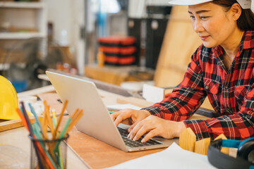 Carpenter woman in a white hardhat and red plaid shirt uses a laptop in a woodshop, surrounded by tools, wood scraps, and pencils. Ideal for online learning, client orders, or woodworking projects