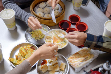 Top view of friends sharing a traditional Taiwanese meal, including xiao long bao, savory pancakes, and congee. A vibrant display of local delicacies served on stainless steel plates