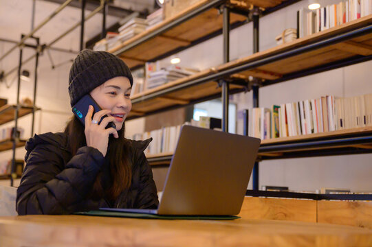 A young Asian woman in winter attire working on a laptop and talking on the phone in a cozy cafe, remote work, or freelance lifestyle.