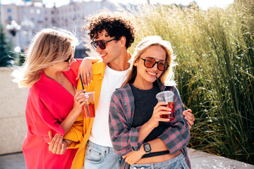 Young three stylish friends posing in the street. Fashion man and two cute female dressed in casual summer clothes. Smiling models having fun. Cheerful women and guy outdoors, Hold and drink lemonade