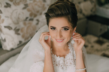 A woman is wearing a white dress and is smiling. She is wearing a pair of earrings and is touching her ears