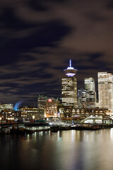 Obraz premium Night view of Vancouver Lookout Tower with illuminated observation deck against urban skyline, featuring waterfront buildings and port infrastructure along harbour