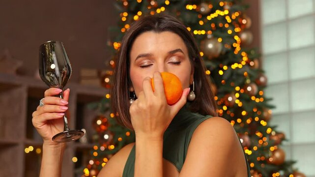 attractive woman sitting against decorated christmas fir tree in front of table and pretend to eat tangerine with fork and knife.girl in gress celebrate winter holidays,new year.cozy home interior