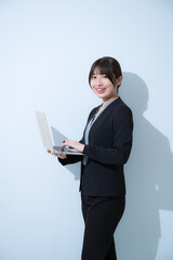 水色背景でスーツを着てカメラ目線のノートパソコンを開く、持つビジネスイメージの爽やかな若い女性　縦