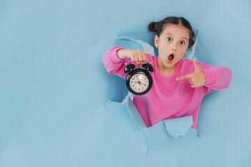 Surprised young girl pointing at alarm clock held in hand, emerging from torn blue paper...