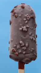 Detailed close-up of a chocolate-covered ice cream bar with a frosty texture, sprinkled with nuts, set against a vibrant blue background.