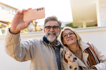 Happy senior couple taking a selfie outside a modern house, smiling at the camera on a sunny day.