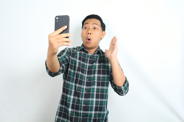 surprised asian man looking at smartphone with shocked face, wow expression, and raising hands, isolated on white background