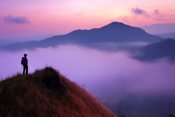 Obraz premium Lone hiker overlooking misty mountains at dawn, basking in the soft glow of sunrise with vibrant hues in the sky and serene landscape below.