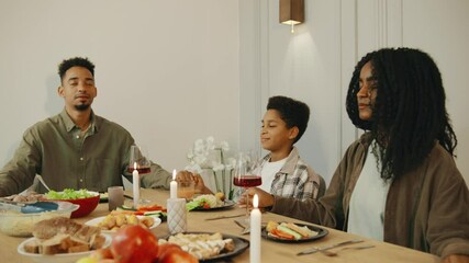 An African American family holds hands and closes their eyes as the father leads a heartfelt prayer of gratitude before their meal, sharing a moment of faith and unity.