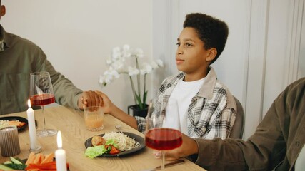 An African American family holds hands and closes their eyes as the father leads a heartfelt prayer of gratitude before their meal, sharing a moment of faith and unity.