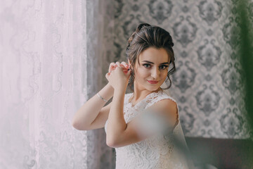 Fototapeta premium A woman is getting ready for a wedding, putting on her wedding dress and earrings. She is standing in front of a window, and the room has a vintage feel to it