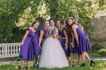 A group of women are posing for a photo in front of a tree. One of the women is wearing a white dress and holding a bouquet of flowers. The other women are wearing purple dresses