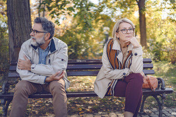 Senior couple sitting apart on a park bench in an autumn setting, looking upset and distant,...