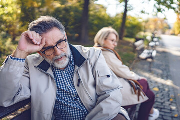 Senior couple sitting back-to-back on a park bench, looking upset and distant, depicting...