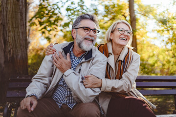 Happy senior couple sitting on a park bench in autumn, smiling and enjoying a sunny day outdoors with colorful leaves in the background.