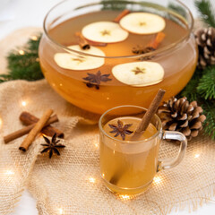 A cup of hot amber apple punch with a cinnamon stick surrounded by fir branches and pine cones with...